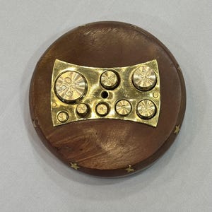 May include: A round, wooden object with a brass plate and weights. The wooden base is brown with small gold star accents around the edge. The brass plate holds several small, circular weights, each with markings. A vintage scale weight set.