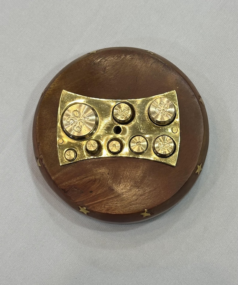 May include: A round, wooden object with a brass plate and weights. The wooden base is brown with small gold star accents around the edge. The brass plate holds several small, circular weights, each with markings. A vintage scale weight set.