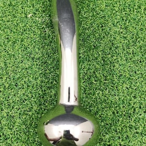 May include: A shiny, chrome-colored metal object with a rounded base and a long, curved handle. The object reflects the surrounding environment, suggesting a polished surface. The item is set against a backdrop of green artificial turf.