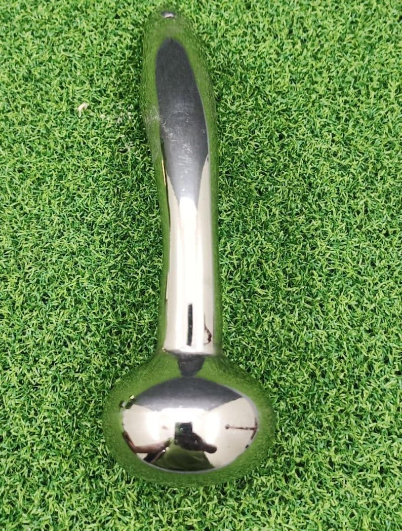May include: A shiny, chrome-colored metal object with a rounded base and a long, curved handle. The object reflects the surrounding environment, suggesting a polished surface. The item is set against a backdrop of green artificial turf.