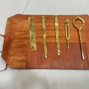 Könnte beinhalten: Ein Satz antiker Zeichenwerkzeuge, darunter ein Lineal, Zirkel und ein Reißzirkel, präsentiert auf einer braunen Leder-Werkzeugrolle. Die Werkzeuge sind aus Messing und silberfarbenem Metall gefertigt. Die Werkzeugrolle besteht aus braunem Leder mit einem Lederriemen.