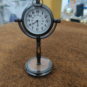 May include: A vintage-style, dark metal desk clock with a white clock face and black numerals. The clock has a round face with a decorative stand and base. The clock's face has the numbers 1 through 24.