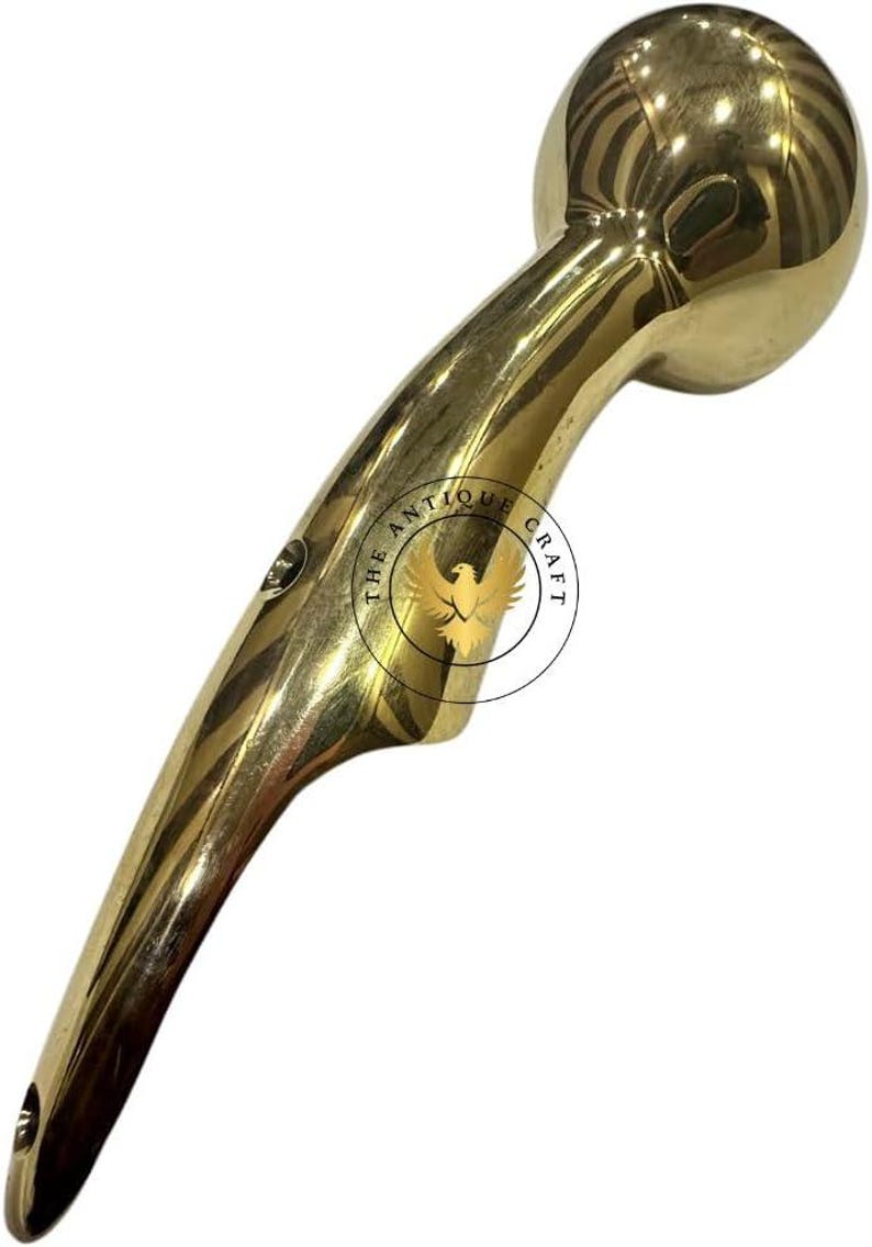 May include: A polished brass handle with a rounded top and a curved, ergonomic design. The handle features a small hole at the base and the text "THE ANTIQUE CRAFT" around a bird emblem. This item is likely a decorative or functional hardware piece.