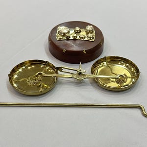 May include: A vintage brass balance scale with two circular pans suspended by chains. A wooden base with brass weights and a brass rod are also present. The scale is likely used for measuring small items or ingredients.