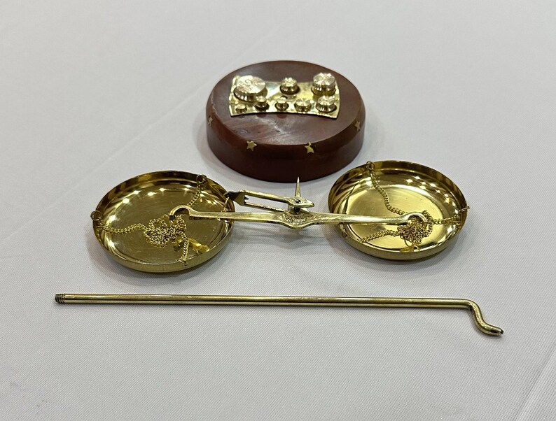 May include: A vintage brass balance scale with two circular pans suspended by chains. A wooden base with brass weights and a brass rod are also present. The scale is likely used for measuring small items or ingredients.