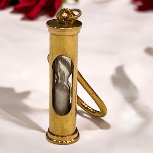 May include: A small, gold-colored hourglass keychain. The hourglass is encased in a cylindrical frame with a loop at the top and a curved handle. The glass portion of the hourglass is visible through an oval opening, and the base has a decorative ring.
