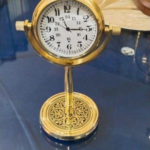 May include: A gold-coloured, vintage-style clock with a white face and black numerals. The clock has a decorative base and a handle at the top. The clock's face displays the numbers 1 to 24.