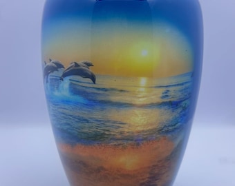 Könnte beinhalten: Blaue Urne mit Sonnenuntergangs- und Delfin-Design. Die Urne hat eine glänzende Oberfläche und einen Deckel. Das Bild zeigt Delfine, die aus dem Meer springen, mit einem Sonnenuntergang im Hintergrund. Der Boden der Urne ist orange und braun.