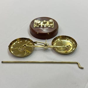 May include: A vintage brass balance scale with two round pans suspended by chains. A wooden base with brass weights and a long brass rod are also present. The scale is likely used for measuring small items or ingredients.