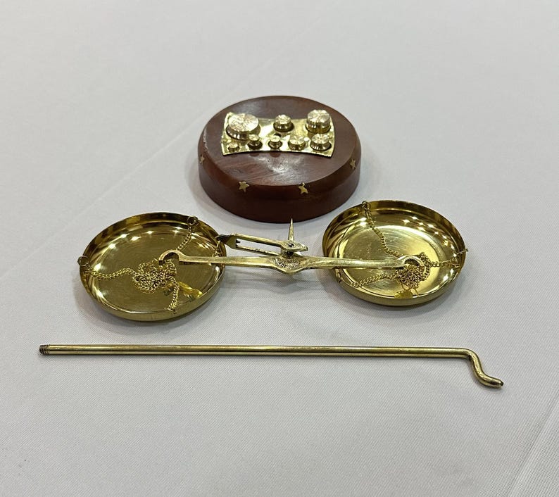 May include: A vintage brass balance scale with two round pans suspended by chains. A wooden base with brass weights and a long brass rod are also present. The scale is likely used for measuring small items or ingredients.