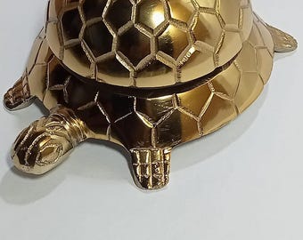 May include: A gold-coloured turtle-shaped bell. The bell has a textured, hexagonal pattern on its shell and a small handle on top. The turtle has detailed feet and a tail. The bell is likely made of metal.