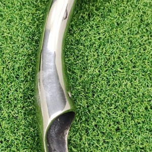 May include: A polished, chrome-like metal object with a rounded top and a curved, elongated handle. The object is set against a backdrop of green artificial turf. The reflective surface of the metal creates highlights and shadows.