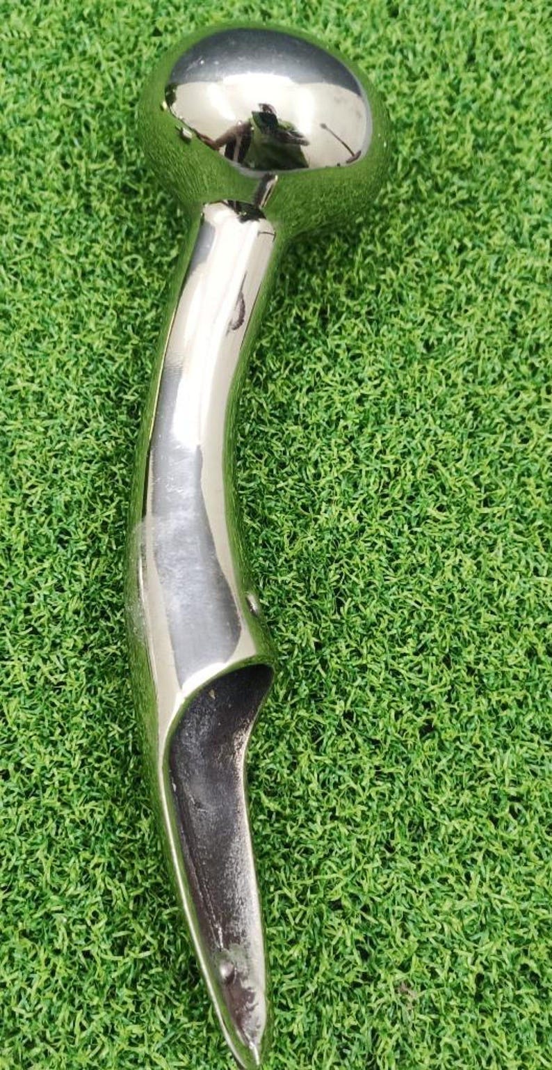 May include: A polished, chrome-like metal object with a rounded top and a curved, elongated handle. The object is set against a backdrop of green artificial turf. The reflective surface of the metal creates highlights and shadows.
