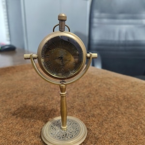 May include: A vintage brass desk clock with a round face and Roman numerals. The clock is supported by a decorative stand. The clock face reads "VICTORIA LONDON". The clock is a classic piece of decor.