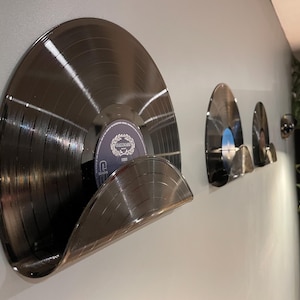 May include: Wall-mounted vinyl record holders made from repurposed records. The holders are a dark metallic colour and are mounted on a white wall. The holders are designed to display vinyl records. The holders are a unique and stylish way to showcase a record collection.