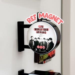 May include: A circular refrigerator magnet featuring a black and white photograph of seven individuals. The text reads BTS WORLD TOUR ARIRANG. The magnet is mounted on a black bracket extending from the surface. The words REF MAGNET appear in pink.
