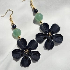 Peut inclure: Une paire de boucles d'oreilles pendantes avec des pendentifs en forme de fleurs noires et des centres dorés. Chaque boucle d'oreille a un crochet doré, des perles noires et dorées et une perle vert clair.