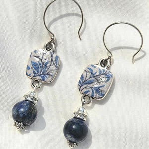 Könnte beinhalten: Ohrringe zum Aufhängen mit silberfarbenen Haken. Jeder Ohrring hat ein quadratisches, silberfarbenes Oberteil mit blau-weißem Blumenmuster. Unterhalb hängt eine dunkelblaue, kugelförmige Perle an einer dekorativen silberfarbenen Kappe.
