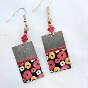 May include: A pair of rectangular earrings with a floral design. The top portion is a dark gray metal, while the bottom features red, yellow, and white flowers on a black background. Gold-colored beads and red crystals accent the gold-tone ear wires.