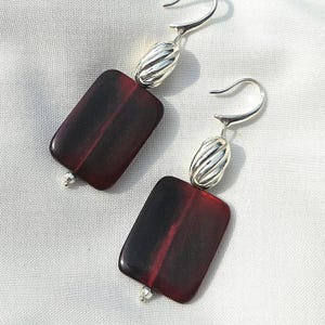 May include: A pair of dangle earrings featuring rectangular, dark red-burgundy stones with a vertical stripe. The earrings have silver-colored, ribbed oval accents and curved ear wires. The earrings are displayed on a white surface.