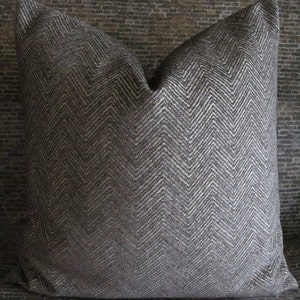 May include: A square decorative pillow with a gray and white chevron pattern. The pillow has a textured appearance and is likely made of fabric. The pillow is placed on a patterned surface.