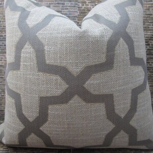 May include: A square decorative pillow with a neutral beige background and a geometric gray pattern. The pillow has a textured appearance and is likely made of a woven fabric. The design features intersecting lines forming a repeating shape.