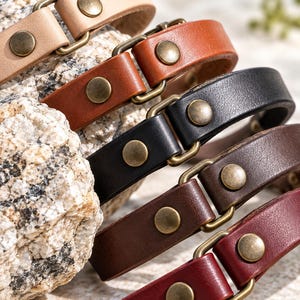 Può includere: Una collezione di cinque bracciali in pelle nei colori beige, cuoio, nero, marrone scuro e bordeaux. Ogni bracciale presenta un cinturino in pelle liscia fissato con rivetti in metallo color ottone anticato e un anello di giunzione rettangolare.