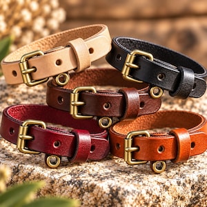 Può includere: Una collezione di cinque bracciali in pelle nei colori beige, nero, marrone scuro, bordeaux e marrone chiaro. Ogni bracciale presenta un cinturino in pelle liscia, una fibbia in metallo color ottone, un passante in pelle e un piccolo anello.