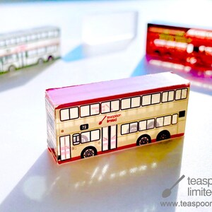 May include: Three miniature double-decker bus models made of clear acrylic. The foreground bus is cream-coloured with red trim, featuring the text "teaspoon limited" and the number "72". Two additional blurred bus models appear in the background.