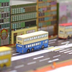 Può includere: Un modello di una strada di Hong Kong con edifici colorati e autobus a due piani. Gli autobus sono gialli e bianchi con accenti blu. Gli edifici sono multicolori e hanno finestre e porte.