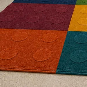 May include: A rectangular area rug featuring a grid of solid color blocks in dark blue, green, yellow, orange, and maroon. Each colored section contains a pattern of raised circular studs, mimicking the appearance of a plastic building block.