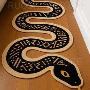 Puede incluir: Una alfombra beige y negra con forma de serpiente y un detallado patrón tribal. La cabeza de la alfombra presenta ojos amarillos y una lengua bífida. La alfombra está colocada sobre un suelo de madera, añadiendo un elemento decorativo único al espacio.