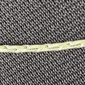 May include: Close-up of a delicate, light-colored chain against a textured, dark gray background. The chain appears to be made of small, interconnected links, suggesting a necklace or bracelet.