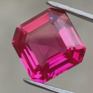 May include: A faceted, vibrant pink gemstone held by tweezers. The gem has a square shape with beveled edges, reflecting light and showcasing its internal structure. The color is a deep, saturated pink, with a hint of red.