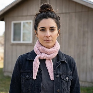 Puede incluir: Bufanda de punto rosa claro anudada en la parte delantera, usada con una chaqueta vaquera oscura. La bufanda está hecha de un hilo grueso y tiene un acabado texturizado. Un edificio de madera en el fondo.