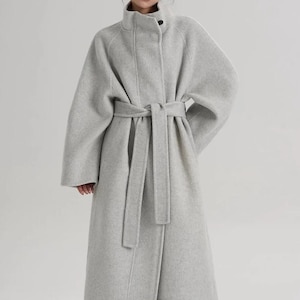 May include: A long, light gray wool-blend coat featuring a high stand-up collar with a single visible button closure, wide sleeves, and a matching fabric tie belt at the waist. The coat has a straight, structured silhouette that falls below the knees.