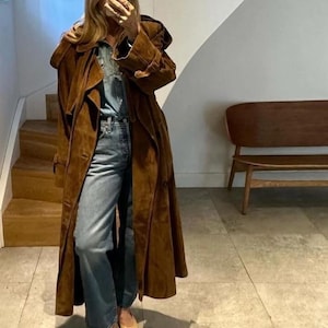 Peut inclure: Un long trench-coat en daim marron avec une capuche. Le manteau est porté sur une chemise en jean et un jean bleu. La personne porte des talons beiges et noirs. La tenue est dans un intérieur aux tons neutres.