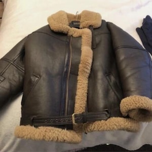 May include: A dark brown leather jacket with a shearling collar and lining. The jacket features a front zipper, a belt at the waist, and shearling trim around the cuffs. The jacket is a classic style, suitable for cold weather.