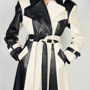 Puede incluir: Un abrigo tipo trench de cuero negro y crema. El abrigo presenta una cintura con cinturón, un cuello con muescas y un diseño geométrico de bloques de color. Las mangas tienen puños negros y el abrigo tiene bolsillos.