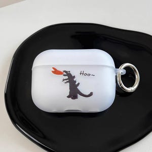 May include: White AirPods case with a cartoon Godzilla design. The design features a black silhouette of Godzilla with orange flames and the word "Hoo~". The case has a silver ring for attachment.