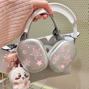 以下が含まれることがあります： ピンクの星のデザインが施されたシルバーのオーバーイヤーヘッドホン。ヘッドホンは白いヘッドバンドとグレーのイヤーカップを備えています。小さなぬいぐるみとピンクのバッグも表示されています。