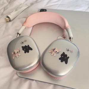 以下が含まれることがあります： 猫と星の装飾が施されたシルバーとピンクのオーバーイヤーヘッドフォン。ヘッドフォンはピンクのヘッドバンドと白いイヤーカップを備えています。イヤーカップは、薄いピンクの猫、黒猫、星型のアクセントで装飾されています。