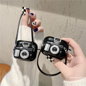May include: Two black AirPod cases designed as vintage cameras, each with a silver lens and flash. They have a black strap and are held up by a person. The cases have the text "OPTICALS" and "1:3.5-5.4" on the lens.