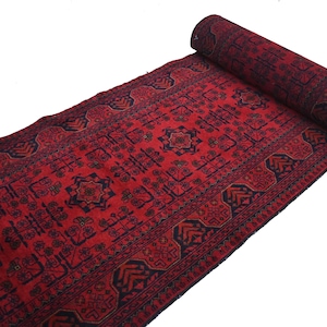 May include: A rolled-up, rectangular red rug with a detailed geometric pattern in dark blue and brown. The rug is partially unrolled, revealing its intricate design and texture. The rug is on a white background.