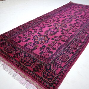 May include: A rectangular, hand-woven rug in a vibrant fuchsia hue. The rug features an intricate, repeating geometric pattern in dark navy and touches of gold. The edges are finished with a dark navy border and white fringe.