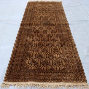May include: A rectangular runner rug featuring a repeating dark brown hexagonal pattern with floral motifs on a lighter brown background. The rug is framed by a decorative border and finished with short, light-colored fringe at both ends.