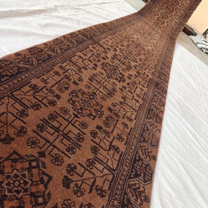 May include: A long, narrow runner rug featuring a deep rust-brown base color with intricate, dark brown geometric and floral patterns. The design includes a central field of repeating motifs framed by a detailed border with repeating shapes.