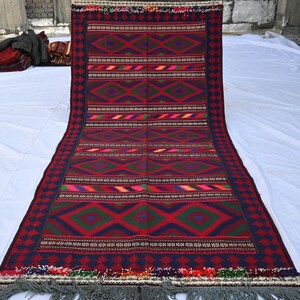 May include: A rectangular, hand-woven rug with a geometric pattern. The rug features a deep red background with green, blue, and yellow diamond and stripe designs. The border is dark blue with a repeating pattern, and the rug has a gray fringe.