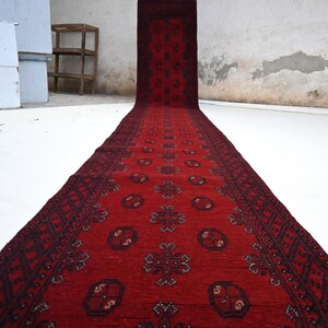 May include: A long, red runner rug with a detailed geometric pattern. The rug features a central design with repeating floral motifs and a dark border. The rug is displayed on a white surface, showcasing its length and intricate design.