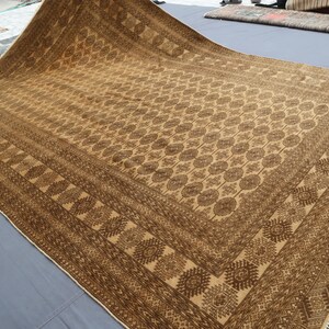 May include: A large rectangular area rug featuring a tan and dark brown color palette. The rug displays a central field of repeating circular geometric motifs surrounded by a wide border with intricate diamond and linear patterns and fringe.